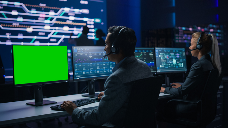 Customer Support Specialist Talks to Client on Headset Works on Green Chroma Key Screen Computer in Big Infrastructure Control Room. Specialists Use Computers Showing Graphs. Talk to Investors