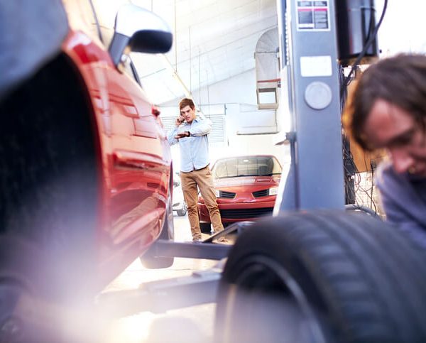 Automotive Service Technician looks at the tire of a car while the client looks at his hand watch.