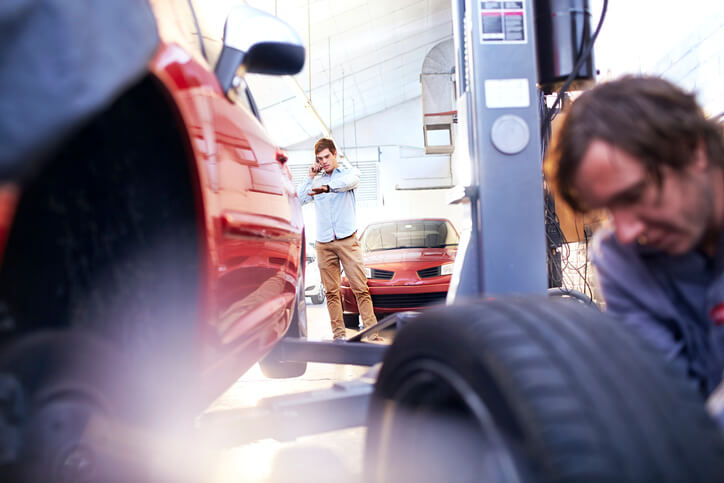 Automotive Service Technician looks at the tire of a car while the client looks at his hand watch.