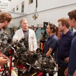 An instructor teaching a group of students about car engines in an auto mechanic training classroom.