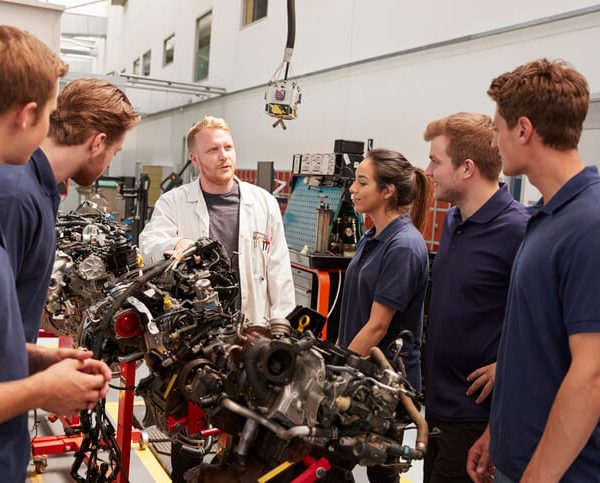 An instructor teaching a group of students about car engines in an auto mechanic training classroom.