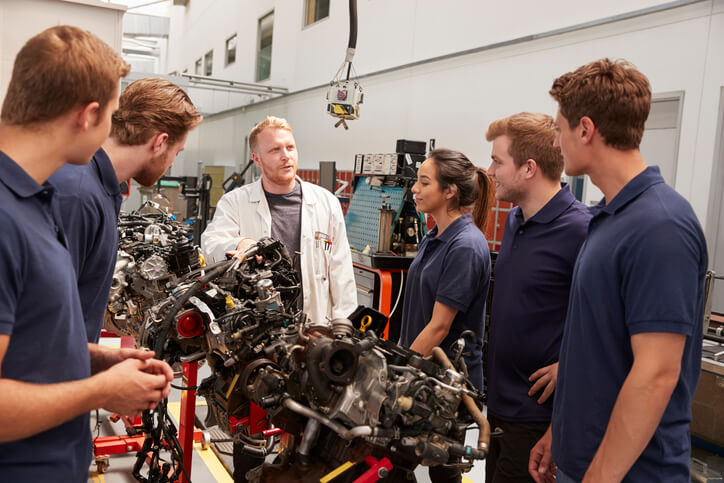 An instructor teaching a group of students about car engines in an auto mechanic training classroom.