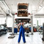 An auto mechanic training student adjusting posture while working under a raised vehicle in the shop