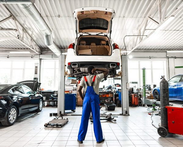 An auto mechanic training student adjusting posture while working under a raised vehicle in the shop