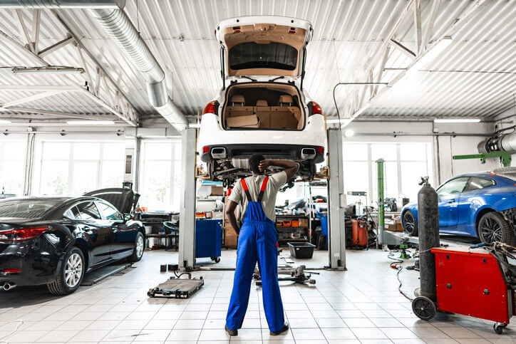 An auto mechanic training student adjusting posture while working under a raised vehicle in the shop