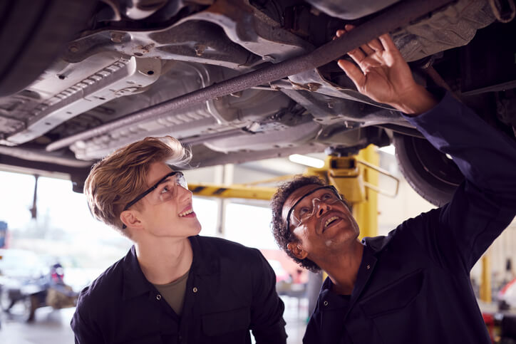A student in auto mechanic training working on an engine with an instructor nearby, highlighting a learning moment.