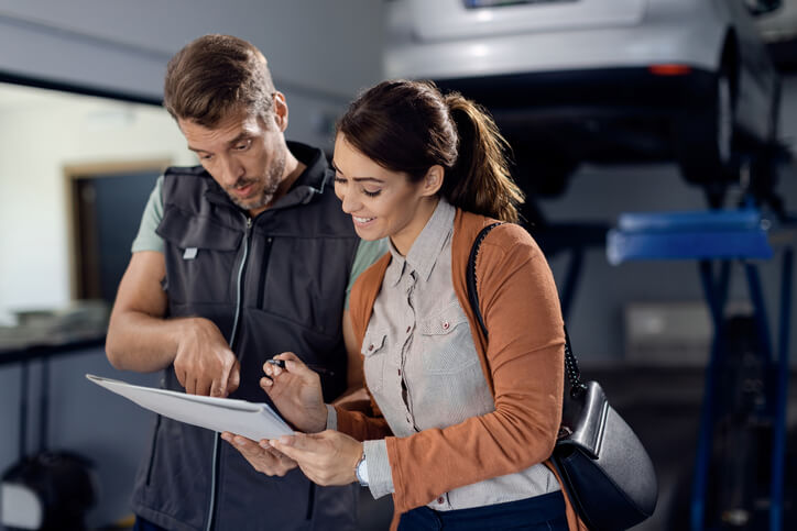 An auto mechanic training graduate and a customer review a repair report and service paperwork in an auto shop.
