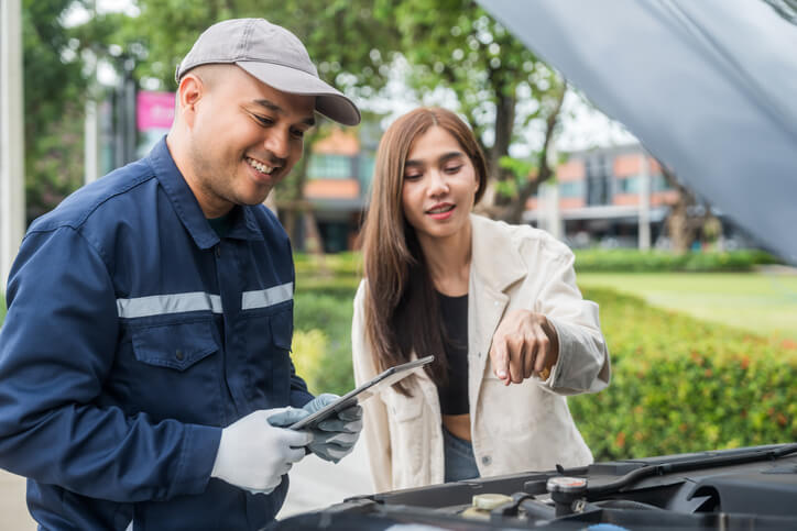 Over the vehicle, an auto mechanic training graduate reviews a clear repair estimate with a customer, explaining costs and recommended work before approval.
