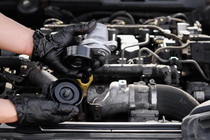An automotive technician inspecting a serpentine belt tensioner in an engine compartment